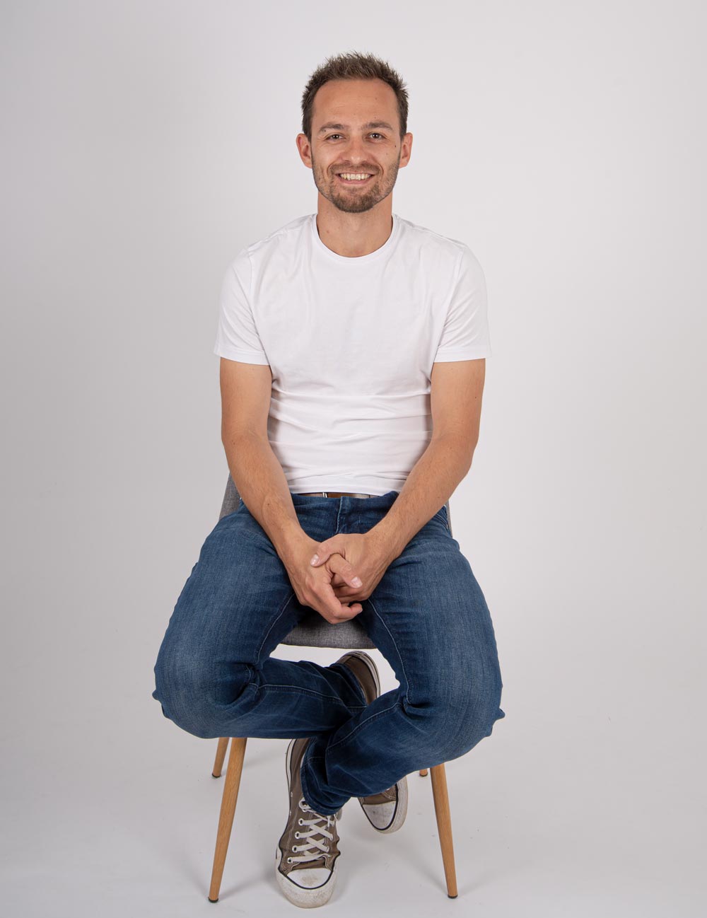 Christoph Bolda sitzt lächelnd auf einem Stuhl in weißem Shirt und in Jeans